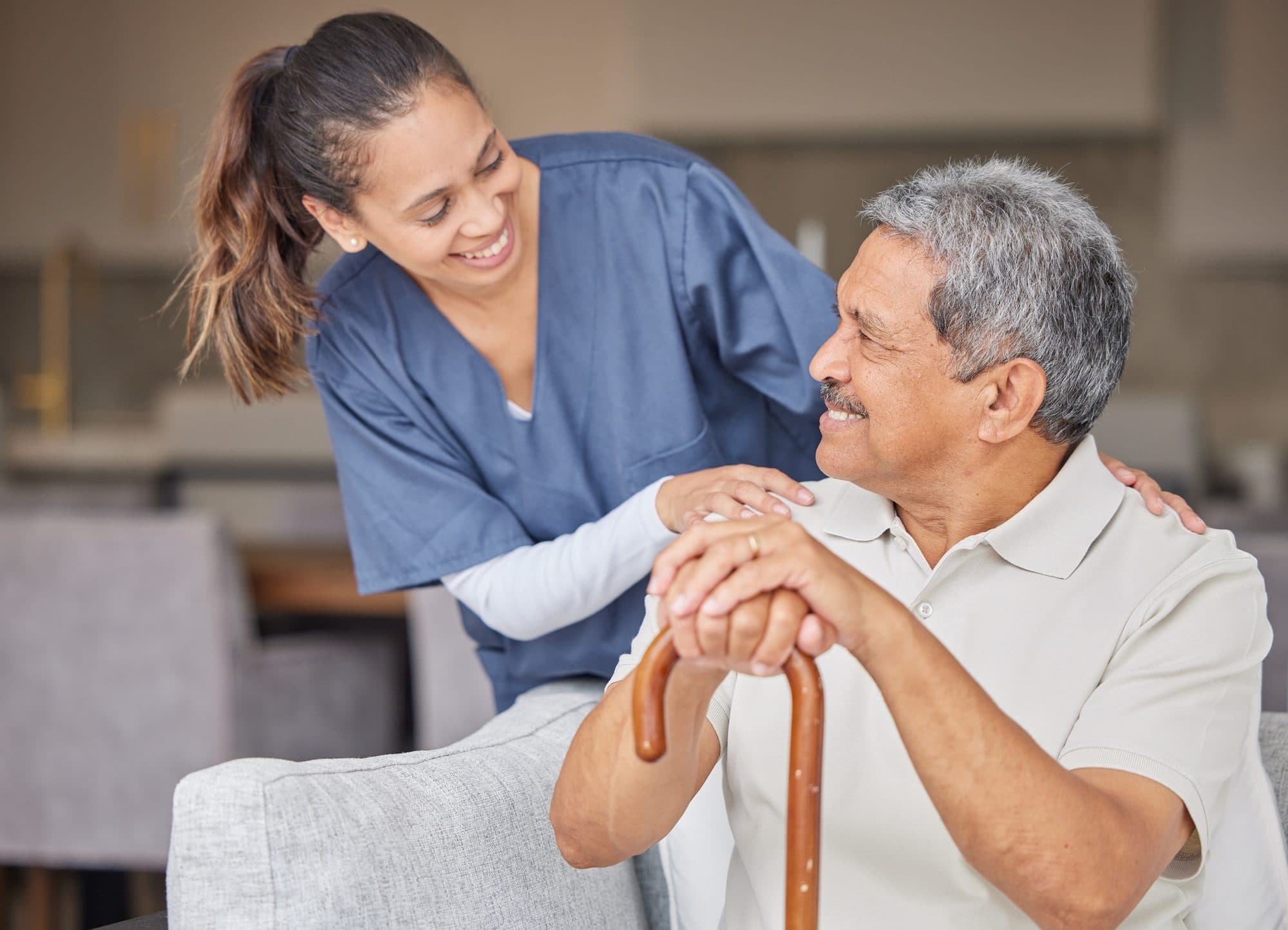 A smiling carer with her hands on the shoulders of an older gentleman holding a walking cane
