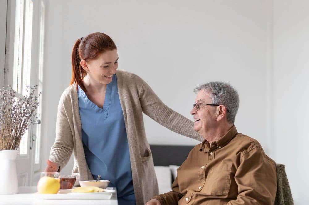A smiling carer resting a hand on the shoulder of an older man at the table